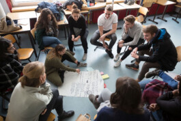 Eine Person schreibt auf ein Flipchart in der Mitte eines Sitzkreises.