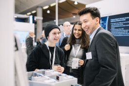 Besucherinnen des Messestands lachen im Gespräch mit einem Mitarbeiter. 