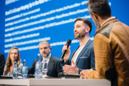 Eine Person spricht auf dem Podium bei einem Vortrag, während andere gespannt zuhören.